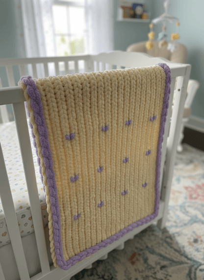 Knitted blanket with purple trim draped over a crib in a nursery.
